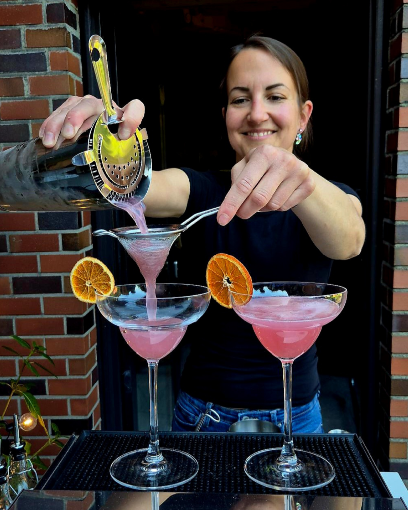 Barkeeperin schenkt auf einer Veranstaltung vom Cocktail Catering der Break Pop-Up Bar pinke Drinks in mit Orangenscheiben garnierte Cocktailgläser ein.